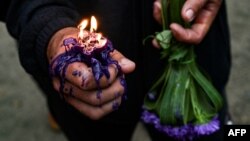 Un devoto sostiene una vela encendida en su mano y un ramo de flores moradas, en tributo a San Lázaro. 