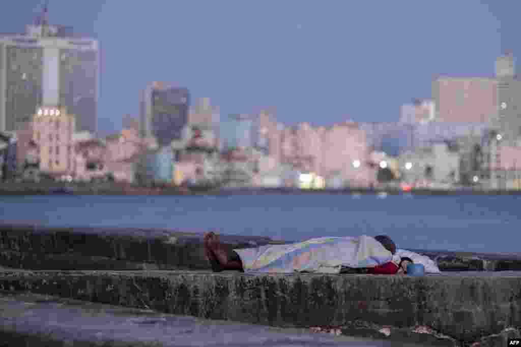 Una persona duerme envuelta en una sábana sobre el muro del malecón al amanecer en La Habana, el 13 de agosto de 2026. YAMIL LAGE / AFP.
