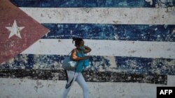 Una mujer camina en la calle. Detrás de ella hay una pared con la bandera de Cuba pintada. (YAMIL LAGE / AFP)
