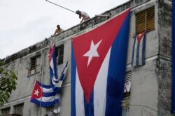Oficialistas cuelgan banderas cubanas en las ventanas de la casa de Yunior García Aguilera, en un intento de evitar que se comunique con el exterior, mientras .sostiene una flor desde una ventana