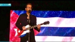 El artista cubano Willy Chirino cantando en un concierto con la bandera cubana detrás de él