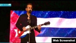 El artista cubano Willy Chirino cantando en un concierto con la bandera cubana detrás de él