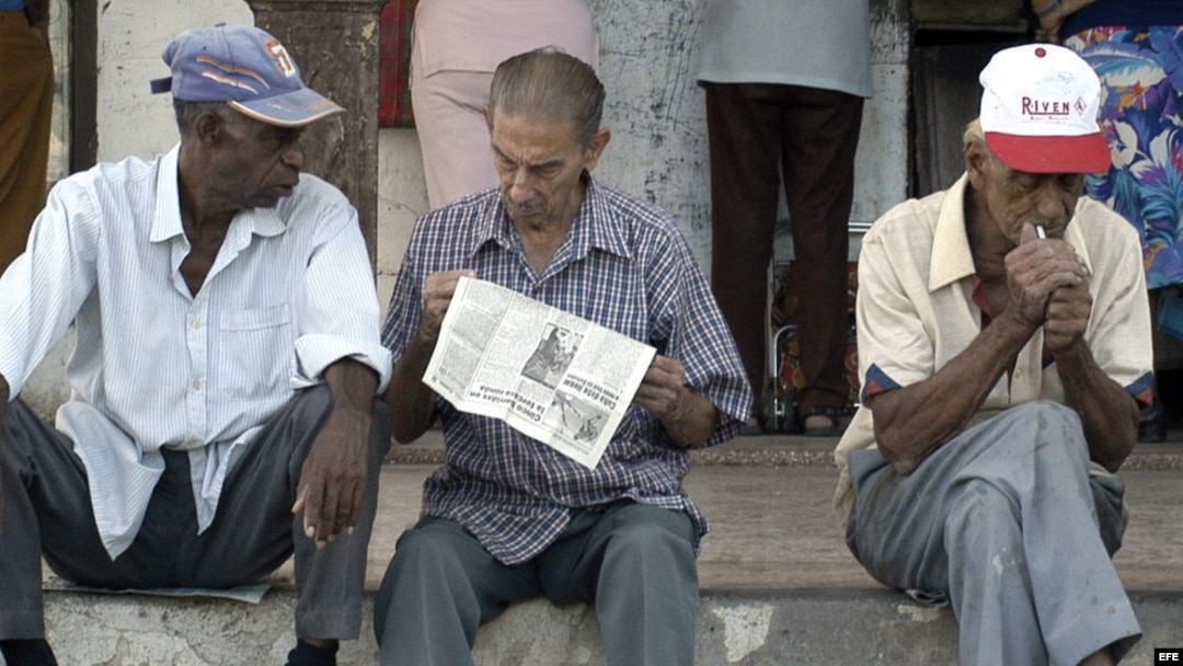 Un grupo de ancianos conversan sentados en un muro mientras esperan la llegada del periódico.