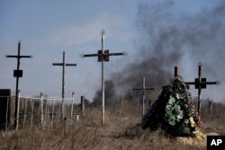 El humo de los proyectiles se eleva al fondo, con una corona de flores en el cementerio de Vasylkiv, el suroeste de Kiev, Ucrania, el sábado 12 de marzo de 2022. (AP Foto/Vadim Ghirda)